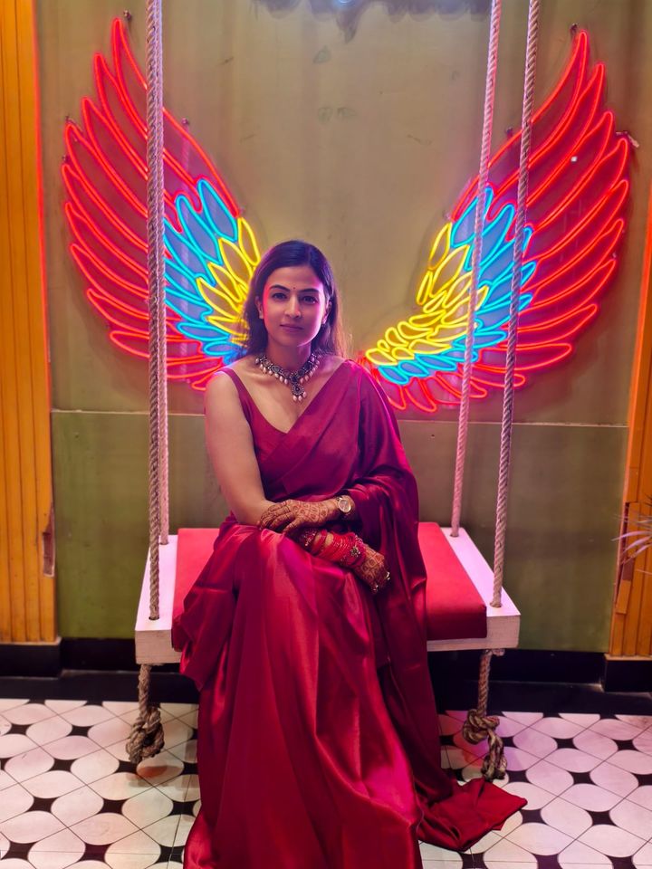 Jyoti in a red saree, seated on a swing in front of colourful neon wings