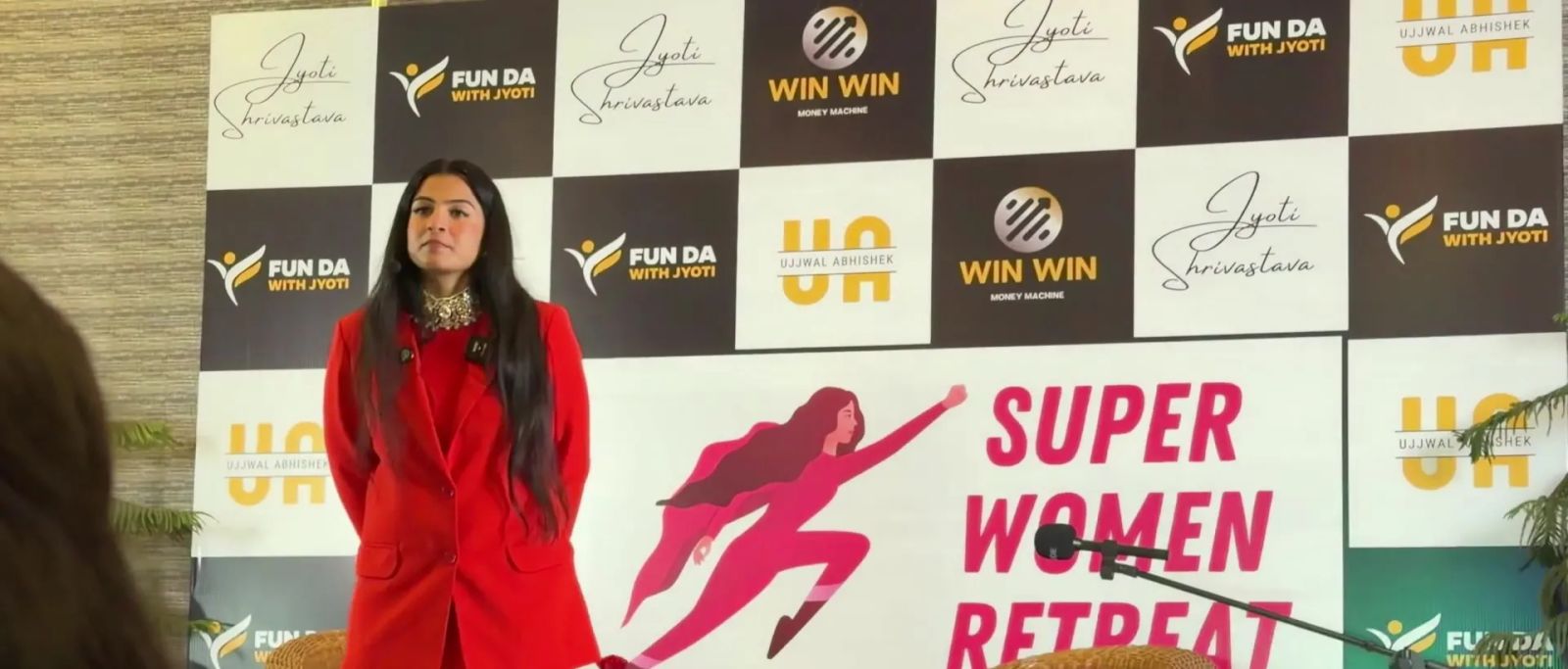 Dr. Jyoti in a red suit on stage in front of the Super Women Retreat backdrop